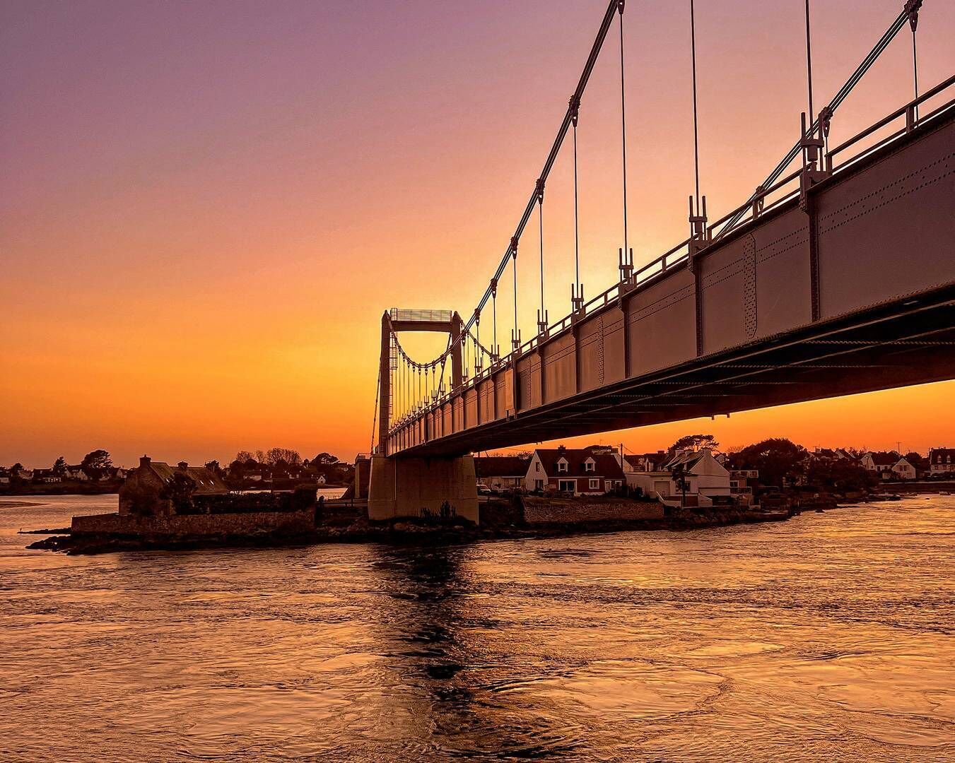Een brug met een hangconstructie, over het water, met huizen op de achtergrond.