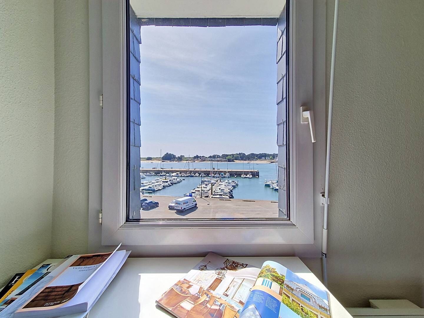Ein Fenster mit Blick auf einen Yachthafen, ein weißer Tisch mit Zeitschriften und eine Wand mit strukturierter Oberfläche.