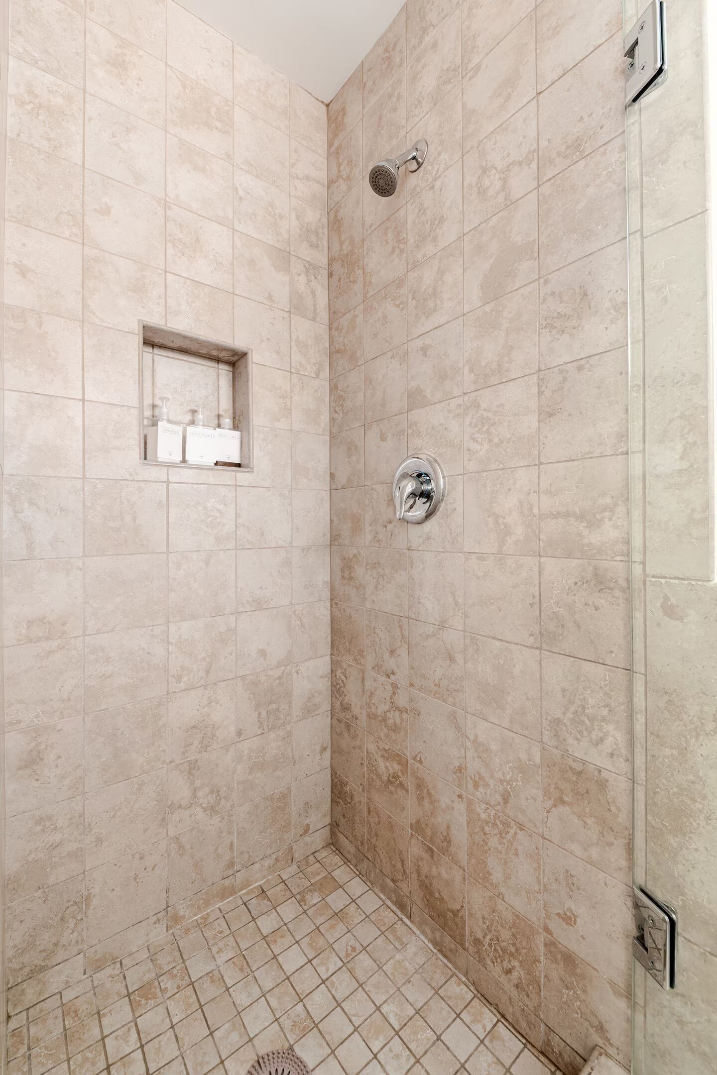 A shower with beige tiles, a glass door, and a small shelf with toiletries.