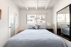 A bedroom with a bed, a nightstand, a mirror, and a window with a view of buildings.