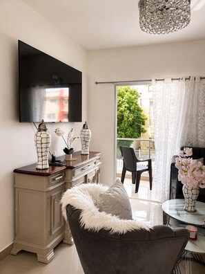 Un salon doté d’un téléviseur à écran plat, d’un meuble décoratif, d’un canapé agrémenté d’un plaid moelleux, d’une table ronde avec un vase, et d’un balcon offrant une vue sur la verdure et les bâtiments.