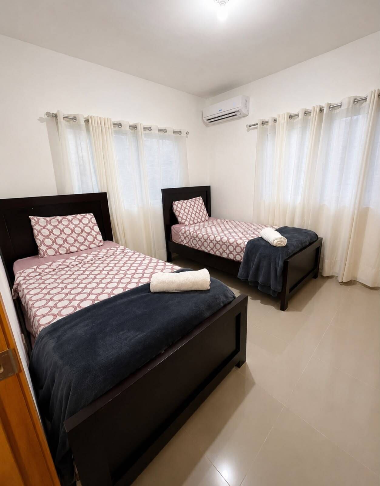 A hotel room with two beds, a wardrobe, an air conditioning unit, and a window with curtains.