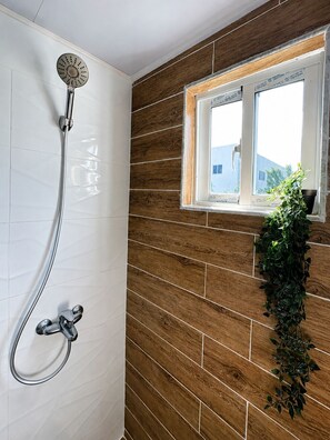 Une douche avec un pommeau de douche Ă effet pluie, une fenĂȘtre donnant sur des immeubles et un mur en bois.