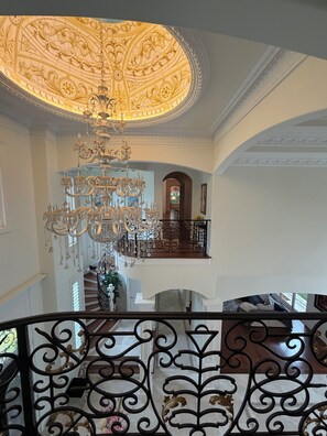 Um interior imponente com um grande candelabro ornamentado, uma escadaria e um balcão com grades de ferro forjado.