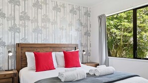 Ein ordentlich bezogenes Bett mit weißen und roten Kissen, einer grauen Decke und aufgerollten Handtüchern. Zwei Nachttischlampen, ein Holz-Kopfteil und ein Fenster mit Vorhängen sind zu sehen.