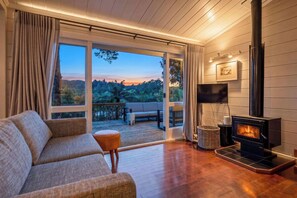 A cozy living room with a sofa, a fireplace, a TV, and a balcony with a view of the forest.