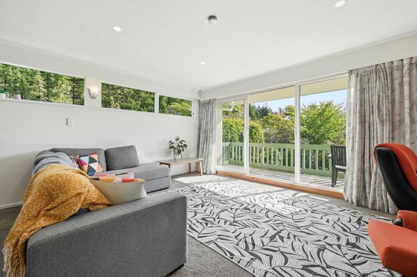 A modern living room with a grey sofa, a red chair, a patterned rug, and a balcony with a view of greenery.