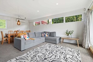 A modern living room with a grey sectional sofa, a wooden dining table with chairs, a patterned rug, and a window with curtains.
