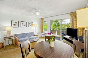 A living room with a sofa, a dining table with chairs, a TV, and a ceiling fan. - Lanai + Pool View Tropical Oasis, Plantation Hale (Kapaʻa)
