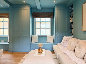 Ein Zimmer mit blauer Wand, einer Fensterbank mit Kissen, einem beigen Sofa und einem Bücherregal.