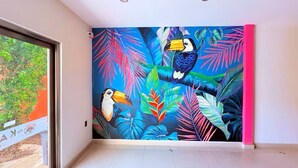 A room with a vibrant mural of toucans and tropical leaves on the wall, a large window, and a tiled floor. - Beautiful King-Size Studio with Panoramic View (Playa del Carmen,)