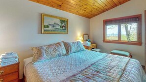 A bedroom with a wooden ceiling, a bed with a quilt, a nightstand, a dresser, a painting on the wall, and a window with a view.