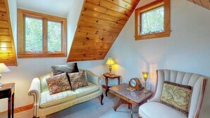 A cozy loft-style room with a sloped ceiling, two windows, a sofa, a chair, a small table, and a lamp.