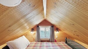 A cozy attic bedroom with a plaid bedspread, wooden walls, and a window with curtains. - Lakefront cottage with full kitchen, firepit, gas grill, Wifi, a/c (Greenville)