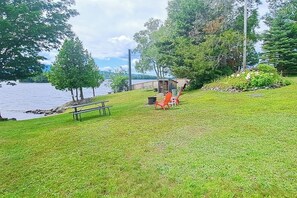 A grassy area with a picnic table and chairs, a small shed, and trees near a body of water.
