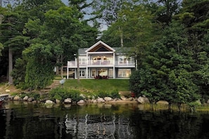 A two-story house with a balcony, surrounded by trees and a body of water. - Elegant lakefront home near Bar Harbor & Acadia with modern kitchen, lake views (Orland)