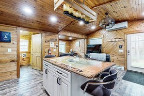 A wooden cabin interior with a kitchen island, dining area, and a large window. - Remodeled Cottage with AC, Private W/D, Full Kitchen, and High-Speed WiFi (Boothbay Harbor)
