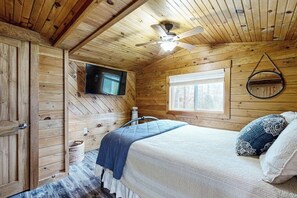 A wooden cabin bedroom with a bed, a ceiling fan, a mounted TV, and a window. - Remodeled Cottage with AC, Private W/D, Full Kitchen, and High-Speed WiFi (Boothbay Harbor)