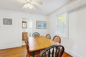 Une salle à manger avec une table et des chaises en bois, un ventilateur de plafond et des tableaux encadrés accrochés aux murs.