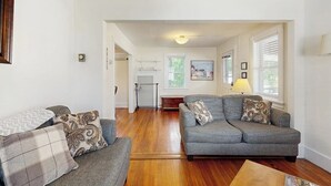 Ein Wohnzimmer mit grauem Sofa, Holzboden, einer Küche im Hintergrund und einem gerahmten Bild an der Wand.