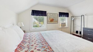 Ein Schlafzimmer mit einem Bett, zwei mit Vorhängen versehenen Fenstern, einer Wand-Klimaanlage, einem gerahmten Bild und einer Kommode.