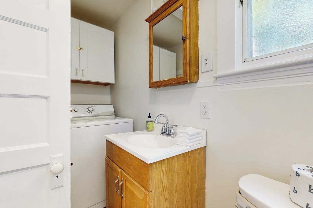 A bathroom with a wooden vanity, a white sink, a mirror, a toilet, and a washing machine.
