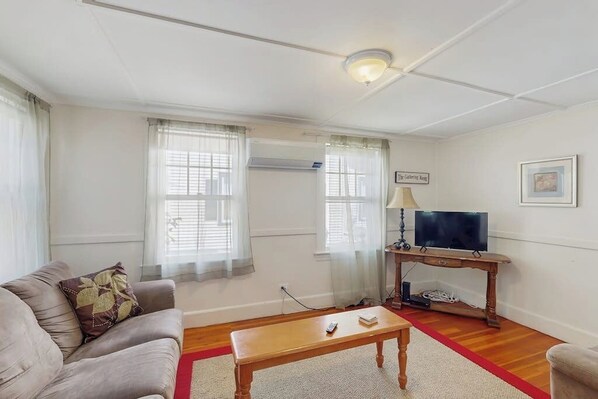A living room with a sofa, a wooden coffee table, a TV on a stand, and a red rug. - Updated, Century-Old Home w/ Glass Porch - Walk into Town/Acadia (Bar Harbor)
