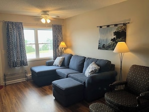 Een woonkamer met een hoekbank, voetenbankje en gestreept fauteuil. Er is een plafondventilator, een raam met gordijnen en een schilderij aan de muur.