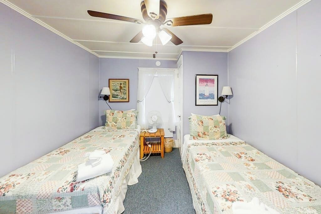 A room with two single beds, a ceiling fan, a small table with a lamp, and a door with a curtain.