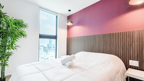 A modern hotel room with a large bed, a potted plant, a view of the city, and a wooden panel wall. - El Semillero (Monterrey)