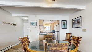 A dining area with a round table and chairs, a kitchen in the background, and a door leading to another room.