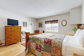 A bedroom with a bed, a wooden dresser, a television on top, a window with a view, and a lamp on a bedside table.