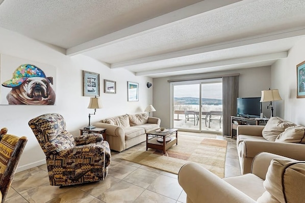 A spacious living room with a sofa, armchairs, a coffee table, and a TV. There is a large painting of a bulldog on the wall, and a balcony with a view.