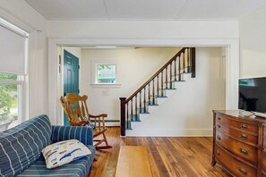 Un salon avec un escalier en bois, un canapé à rayures bleues, un fauteuil à bascule en bois et une commode en bois sur laquelle est posée une télévision.