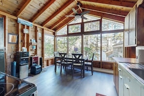 A wooden cabin interior with a dining table, chairs, a ceiling fan, and large windows.