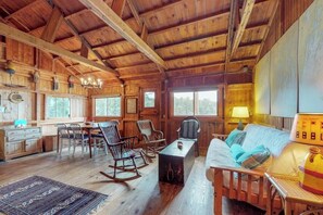 A rustic cabin interior with wooden walls, a dining area, a sofa, rocking chairs, and a lamp.