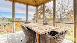 A covered outdoor seating area with a table and chairs, large windows, and a view of trees and landscape.