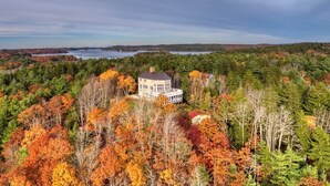 A house surrounded by autumn foliage, with a lake and forest in the background.