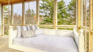 A wooden deck with a bed, large windows, and a view of trees.