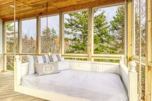A wooden deck with a bed, large windows, and a view of trees.