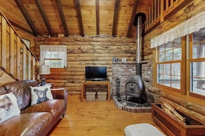 A log cabin interior with a leather sofa, a television, a fireplace, and a window with lace curtains. - Lakefront home w/ pristine views Outdoor firepit and full kitchen (Greenville)