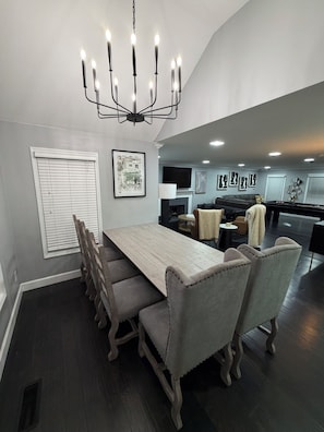 A dining area with a long table, cushioned chairs, a chandelier, and a kitchen in the background.