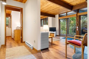 A room with wooden flooring, a desk with a lamp, a chair with a cushion, and large windows with a view of trees. - Beautiful hidden Waterfront Retreat with  Rocky Beach (East Boothbay)