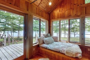 Een chalet met houten inrichting, een bed met kussens, en grote ramen met uitzicht op bos en water.