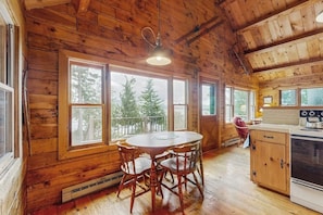 Una cabaña de madera con una mesa y sillas de comedor, una zona de cocina y ventanas amplias que dan a vista de árboles.