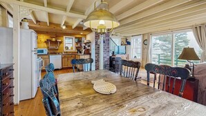 A dining area with a wooden table, chairs, a hanging light fixture, and a kitchen in the background.