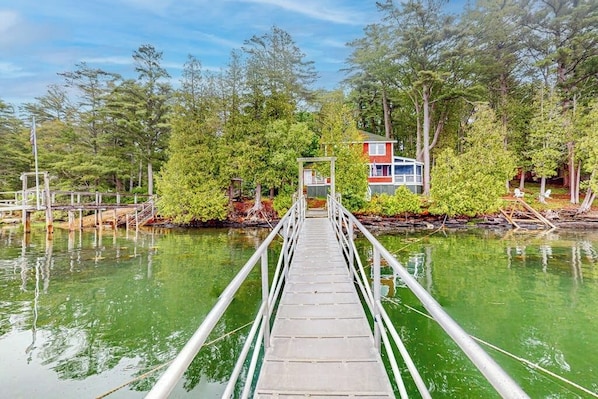 Eine Holzbrücke führt zu einem Haus, das von Bäumen und Wasser umgeben ist.
