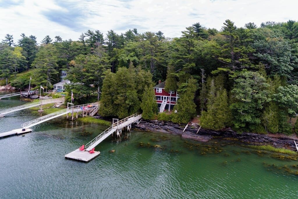 Un muelle que se adentra en un lago, con una casa y árboles al fondo.