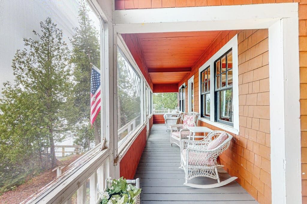 A covered porch with white wicker rocking chairs and a small table, an American flag, and a view of trees.
