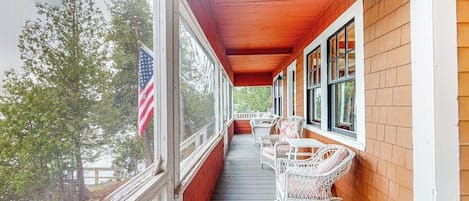 Een overdekte veranda met witte rieten schommelstoelen en een tafeltje, een Amerikaanse vlag, en uitzicht op bomen.